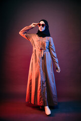 muslim woman wearing black hijab and orange long dress holding sunglasses looking sideways and...
