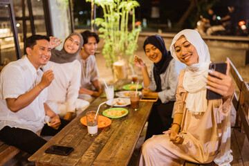 Asian woman in hijab holds cell phone while making video call during iftar with friends at outdoor cafe