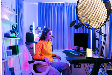 Young Caucasian woman professional gamer wearing yellow hoodie sits on a chair with a gaming table with pc, keyboard, monitor, microphone. Prepare for competition, cast gameplay or record a podcast.