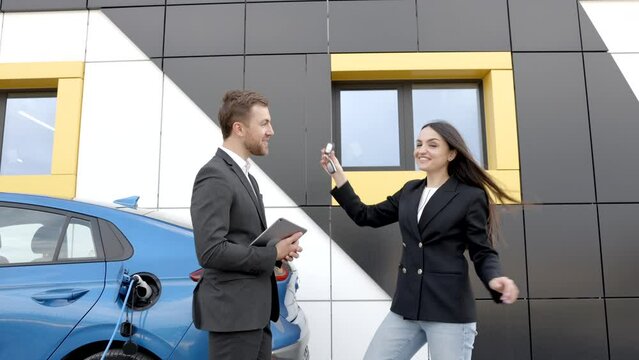 Happy female client standing at brand new car and taking keys from dealer in dealership outdoor. The woman is very happy with the new car, holding the keys in her hands