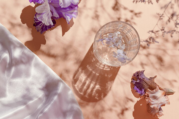 A glass with water and iris flowers on pastel background with shadows and white silk cloth. Summer...