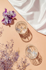 Two glasses with water and iris flower on pastel background with shadows and white silk cloth. Summer refreshment concept. Sunlit flat lay.Top view