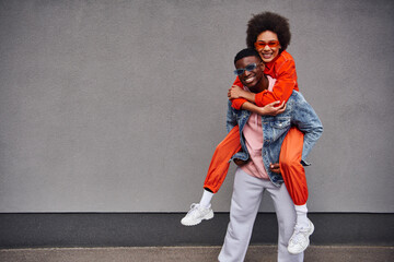 Smiling young african american woman in sunglasses and bright outfit piggybacking on stylish best friend and looking at camera near building on urban street, trendy friends in urban settings