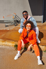 Full length of cheerful young african american woman in sunglasses and bright outfit sitting on...