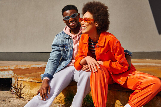 Cheerful young african american best friends in trendy outfits and sunglasses laughing and having fun while sitting on rusty stairs near building on urban street, trendy friends in urban settings