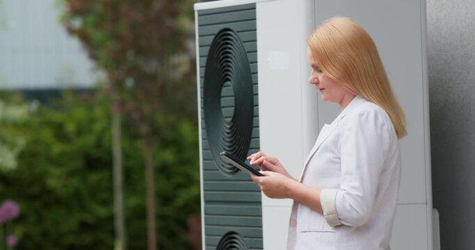 Woman sets up a heat pump near a private house. Uses a tablet