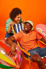 Obraz premium Pleased young african american woman with bold makeup and summer outfit looking at best friend in panama hat sitting on deck chair near ball and swim ring on orange background, fashion-forward friends