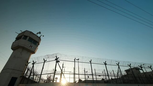 An extreme wide angle of prison and a watchtower, the last sun rays are penetrating through the fence, minutes before sundown.