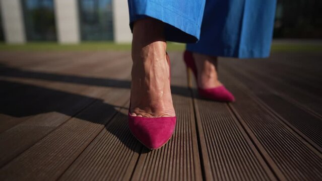 Close-up female foot in red high-heels and blue jeans in sunshine outdoors. Closeup unrecognizable Caucasian woman moving leg in slow motion waiting on urban city street