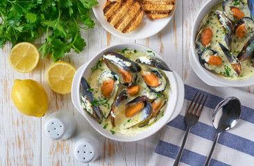 Creamy soup with mussels served with lemon and parsley on a white table, top view.