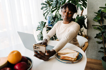 Young black woman using cellphone while working with laptop