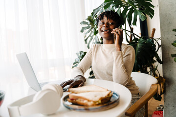 Young black woman talking on cellphone while working with laptop