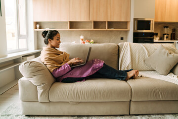 Young indian woman smiling and using laptop while resting on couch