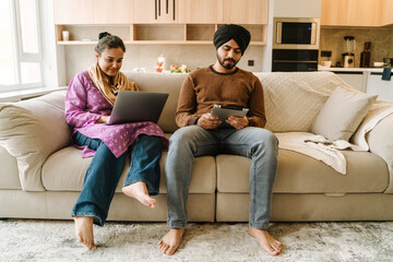 Indian couple sitting on sofa with laptop and tablet