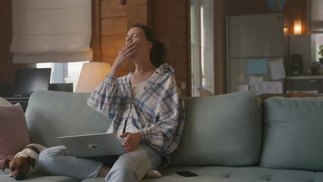 Young woman sitting comfortably on a sofa working on laptop. She is overworked and lets out a tired yawn. Freelancer does monotoneus task, becomes bored. 
