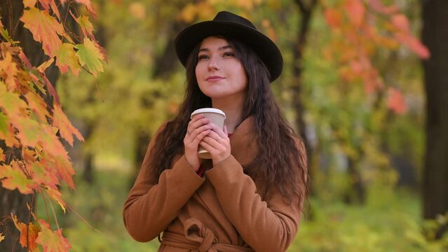 A pretty young woman in a trendy beige coat and hat walks in the park in autumn, admires the yellow leaves and drinks coffee from a disposable cup