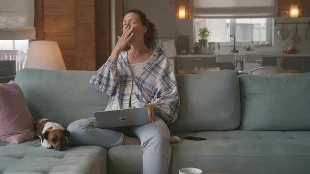 Young woman sitting comfortably on a sofa working on laptop. She is overworked and lets out a tired yawn. Freelancer does monotoneus task, becomes bored. 