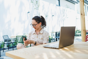 Remote work. Relaxed woman sitting on outdoor terrace in cafe and working online with coffee and phone. Female freelancer works remotely online while sitting in a summer cafe at a laptop