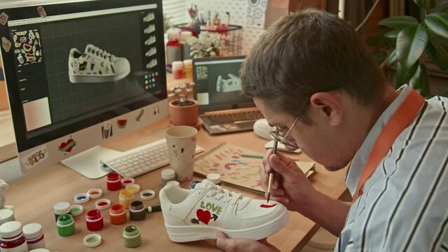 Panning right medium shot of male Caucasian man sitting at desk in loft studio holding white sneaker and drawing picture with red paint on it indoor at daytime