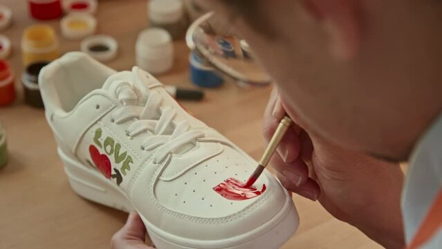 Over-the-shoulder selective focus on hands of male Caucasian man sitting at desk in loft studio holding white sneaker and drawing with red paint on it indoor at daytime