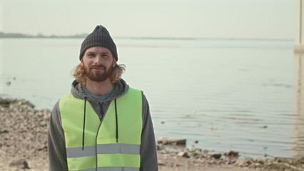 Medium portrait shot of young male environmental volunteer in neon green vest and beanie hat standing on shoreline and posing for camera