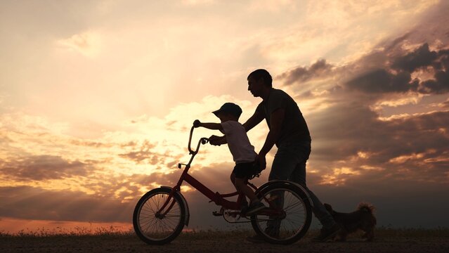 happy family in the park. father teaching son to ride a bike at sunset silhouette in the park. son child learning to ride a bike at sunset father helping son. child playing riding a lifestyle bike