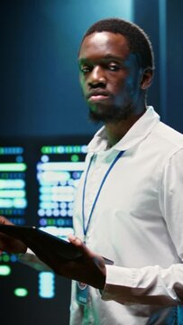 Vertical video African american supervisor in server farm using tablet to find firewall misconfigurations affecting rackmounts performance and connectivity, leading to slow data transfer rates