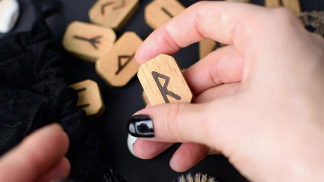 Fortuneteller twists in her hand wooden rune on black table background. Scandinavian magical esoteric signs for divination and prediction of the future and destiny, esotericism concept.