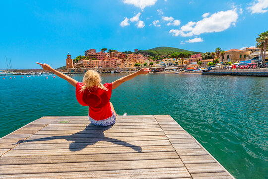 Tourist woman sitting over jetty and looking at Rio Marina harbor of Elba island in red suit. Lifestyle tourist girl on holiday travel in Italy, Europe. Rio Marina skyline cityscape on background,