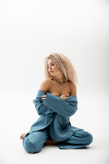 full length of passionate woman with bare shoulders, wavy ash blonde hair and closed eyes sitting in blue oversize suit on grey background, sexy look, fashion and sensuality