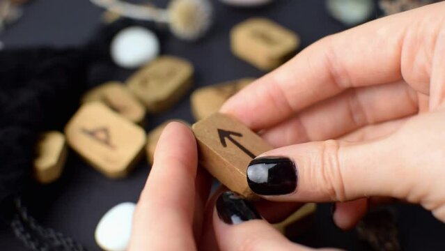 Fortuneteller twists Scandinavian wooden runes in her hands on a black table with a velvet bag and stones. Magical esoteric symbols and signs for divination and prediction of the future and destiny.