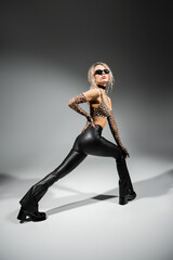 full length of sexy woman in dark sunglasses, animal print crop top, long gloves and black latex pants posing with hand on hip on grey background, glamour style, modern self-expression