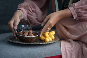 Muslim woman wears hijab sitting on couch and holding dried dates fruit bowl and fresh date nearby in metal tray, iftar meal, Ramadan kareem fasting month concept