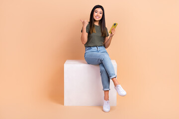 Full body photo of korean young lady wear denim jeans sit box podium point finger with cell phone new website isolated on beige background