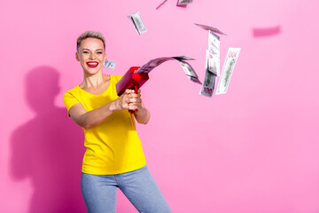 Portrait of overjoyed pretty girl hold money gun shooting dollar bills isolated on pink color background