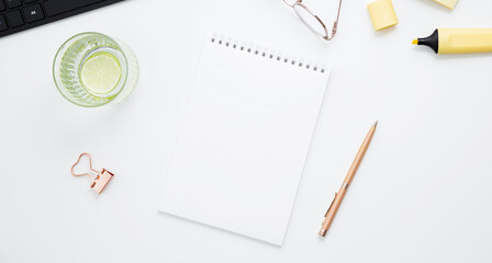 Workplace with white notebook, black keyboard, stationery, glass of water with lime and glasses on white desk. Flat lay office desk, mock up space for text. Top view. Copy space