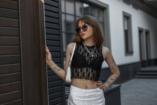 Fashionable beautiful sexy hipster woman model with a tattoo on her arms with vintage sunglasses in a lace black trendy top with a bra and a white skirt with a handbag walks in the city