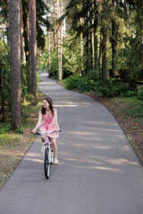 young active woman in pink dress rides a bicycle in park among the trees. Women fun weekend sports and recreation. Walking along bike path in national park, freedom and speed. riding