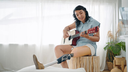 Happy asian woman in leg prosthesis equipment enjoy playing guitar and singing while sitting on...
