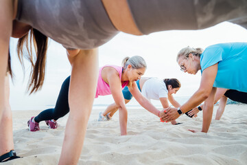 Women of various ages doing fitness workouts in class exercise with coach on beach. Ladies doing paired plank exercises and high-fiving each other. Sport for health and wellbeing. Active lifestyle