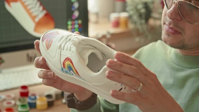 Medium close up of male Caucasian designer examining customized sneakers with colorful drawings indoor at daytime