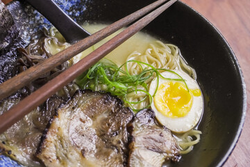 Close-up photo of Japanese pork miso ramen consisting of broth, miso, pork, sliced scallions, sliced mushrooms, soft half boiled egg or ajitsuko tamago ajitama in a bowl on a wooden table.