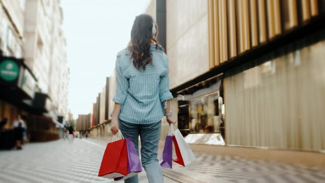Young woman in casual clothes with multicolor paper bags walking near the mall after a successful shopping in a back view. Concept of carefree urban life on a summer sunny day