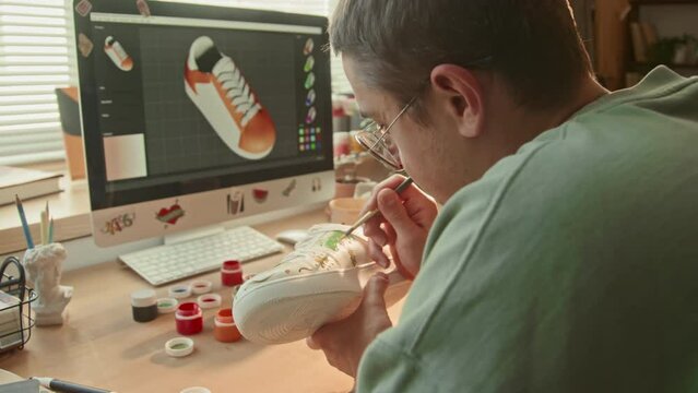 Over-the-shoulder shot of male sneaker designer sitting in front of computer with colorful sketch on screen working on new item