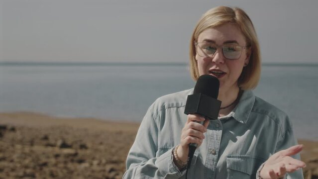 Medium shot of female TV reporter standing on gulf coast on summer day, looking at camera and speaking in mic