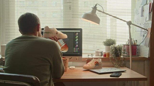 Back view of male sneaker designer sitting in front of computer with colorful sketch on screen customizing new item