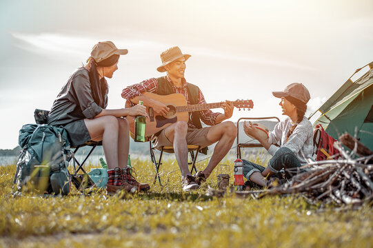 Picnic And Camping Tent, Adventure, Travel, Tourism, Friendship And Recreation Concept. Group Of Asian Tourists Man And Woman Enjoying A Drink After A Set Up Outdoor Tent Playing Music Together.