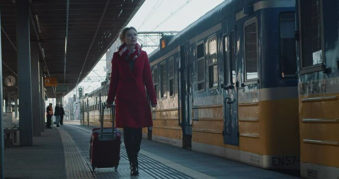 Woman in a Red Coat Traveler with Trolley Bag Walking Along the Station Platform Along the Departing Modern Train. She's About to go on a Train Journey