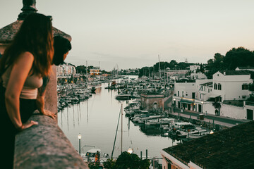 Widok na duży port z jachtami oraz patrzący na niego ludzie o zachodzie słońca na wyspie Menorca - obrazy, fototapety, plakaty