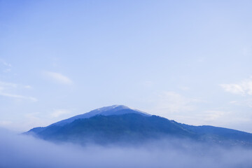 Beautiful view of Mount Merbabu mountainside in the morning with vegetable garden and village settlements or housing in Selo Boyolali, Central Java, Indonesia. Blue sky, fog, mist, clouds backgrounds